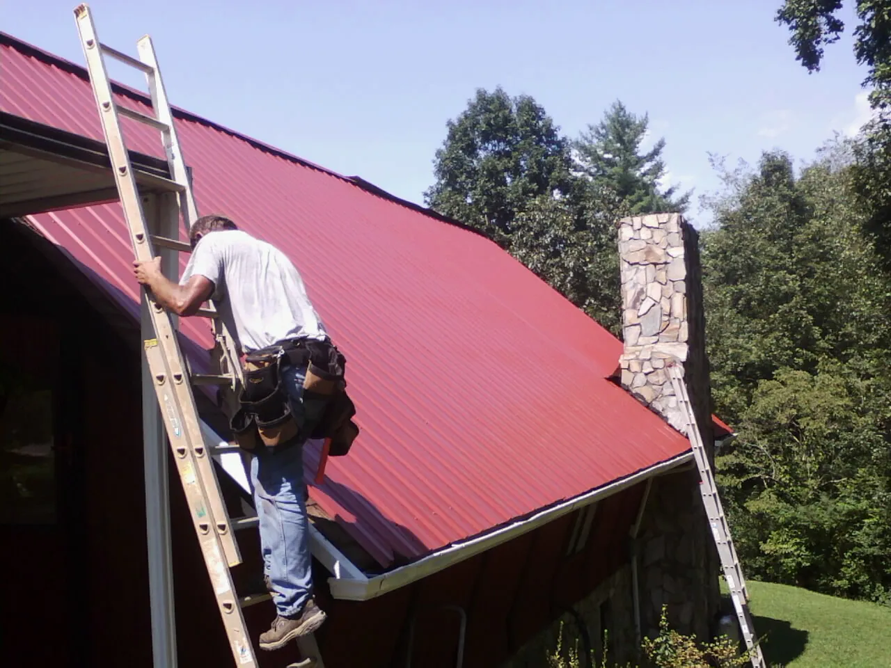 Roofer on a ladder beside a red metal roof and stone chimney — Soffit & Fascia Repair by Roofing Brothers in Coldspring